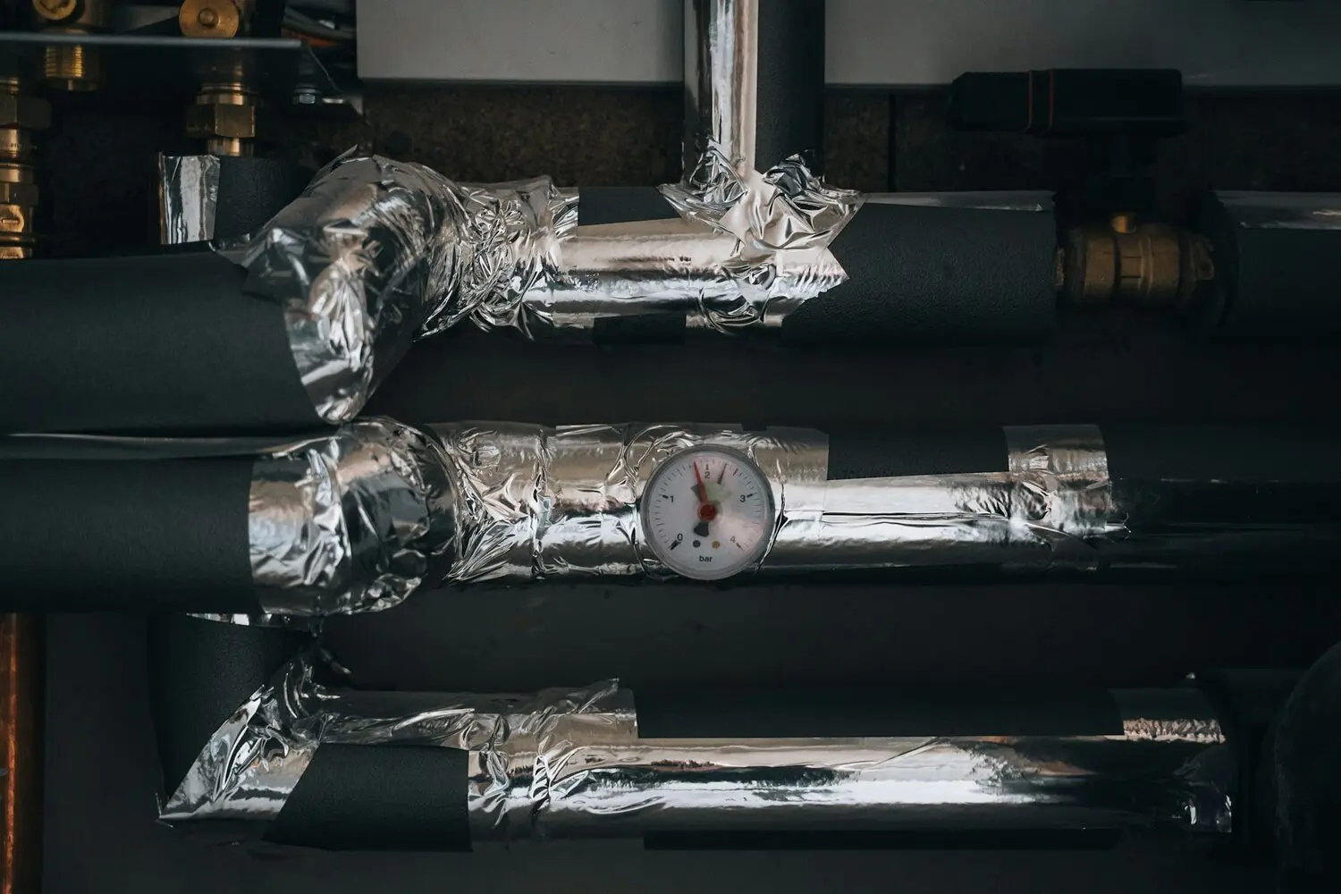 Tuberías de un circuito de agua de climatización forradas con coquilla negra y cinta de aluminio, con un manómetro que marca 2 bar, una válvula de latón y un actuador de válvula de zona